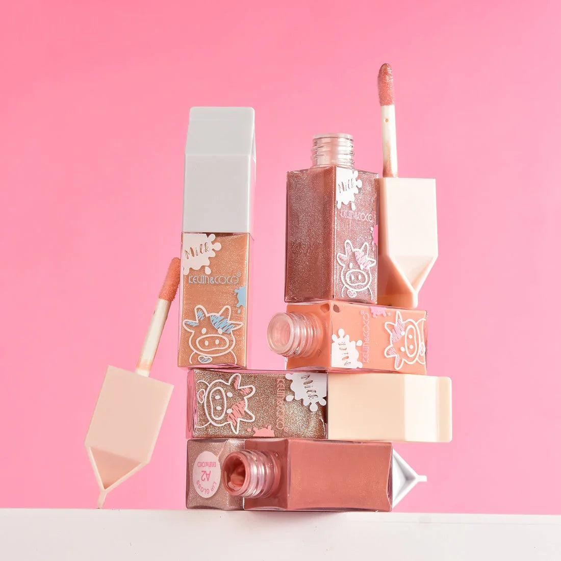 Brillos labiales Bibi Boom en tonos nude y rosa con envases de vaquita, fondo rosa.